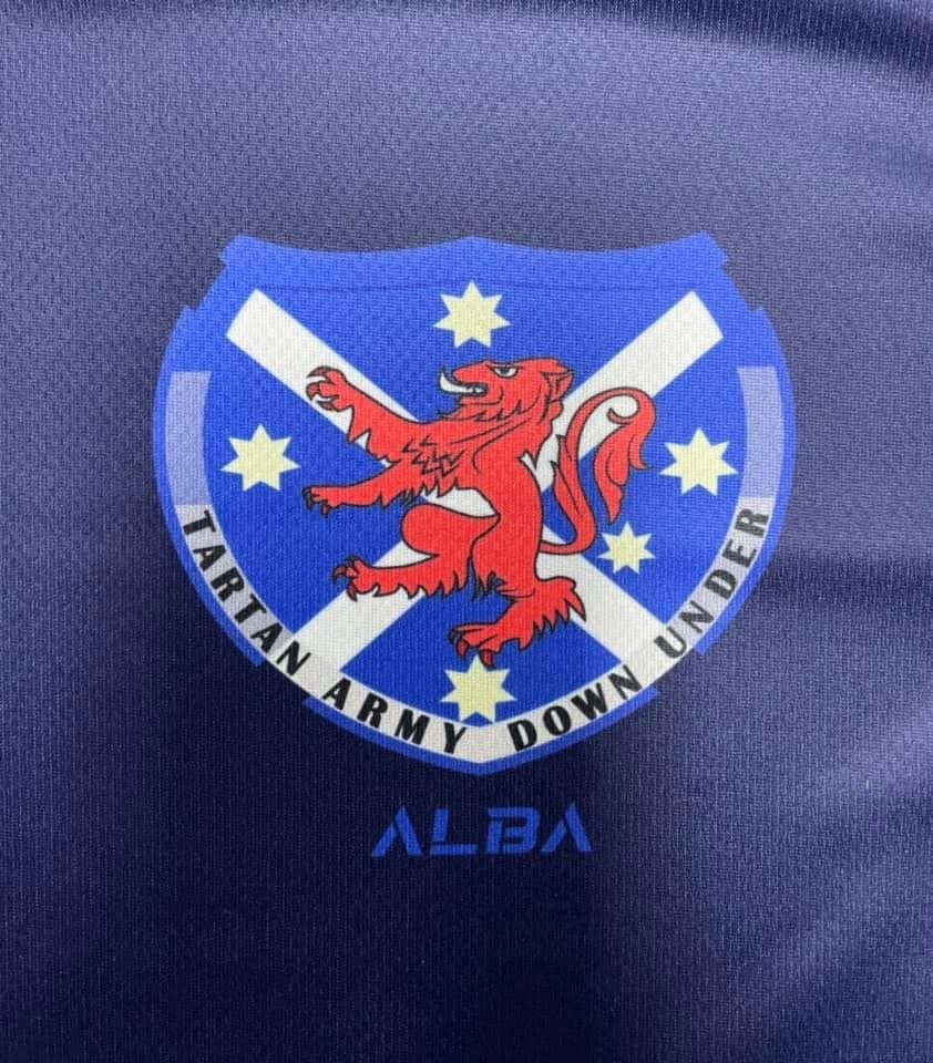 Emblem with a red lion on a blue shield with white cross, stars, and text 'Tartan Army Down Under' on a dark blue background.